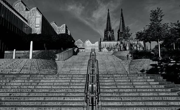 Dramatic black and white image of stairs leading to the Cologne Cathedral, framed by modern architecture and trees under a vivid sky.
