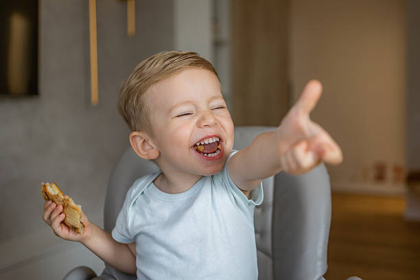 istock Happy young boy laughing and pointing while eating bread at home 2226412464