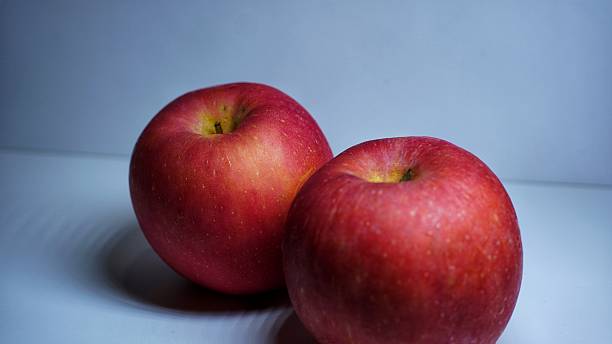 Two fresh red apples placed side by side, showcasing their bright red color and smooth skin texture, against a light gray background with studio lighting.