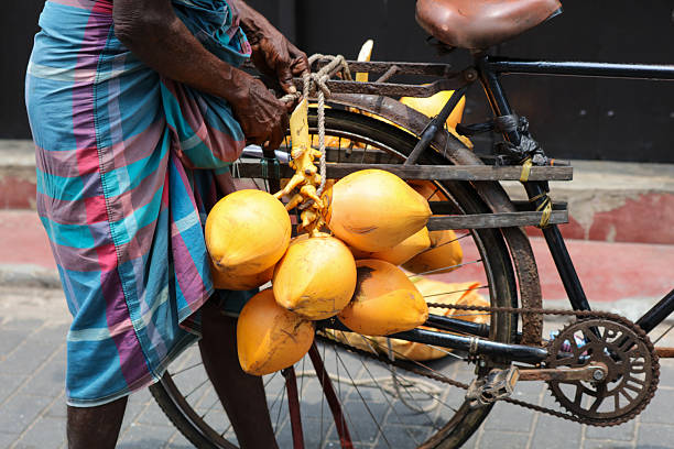 A man is riding a bicycle with a basket full of coconuts. The coconuts are hanging from the basket and the man is holding onto the handlebars. The scene is lively and energetic, with the man