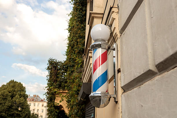 istock Classic barber pole with red, white, and blue spiral stripes mounted on a building wall, with ivy-covered architecture and blue sky in the background 2226419952