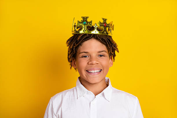 Close-up image of a cheerful boy wearing a gold crown and a white shirt standing in front of a bright yellow background