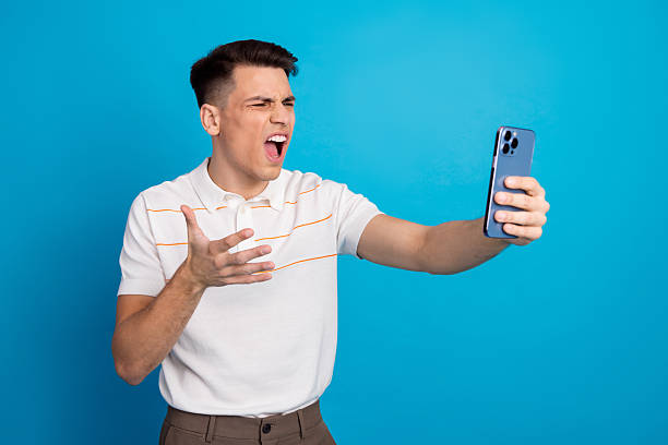 istock Young man angrily gesturing while taking a selfie with a smartphone against a vibrant blue background 2226421782