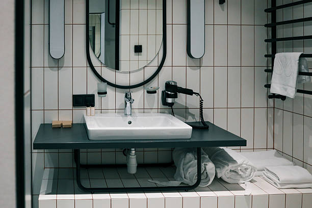 Modern hotel bathroom interior featuring oval mirror, white-tiled walls, sleek countertop basin, chrome faucet, wall-mounted hairdryer and neatly rolled towels for a spa-like ambience