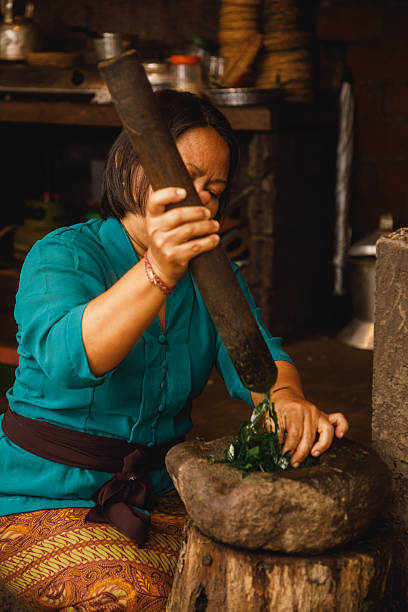 A woman skillfully crushes fresh ingredients in a traditional stone mortar and pestle inside a rustic-style kitchen. This image portrays culinary tradition, cultural heritage, and labor-intensive techniques. The vibrant green herbs and the vintage ambiance evoke a sense of nostalgia and appreciation for homemade preparation methods. Perfect for topics on cooking, tradition, or cultural practices.