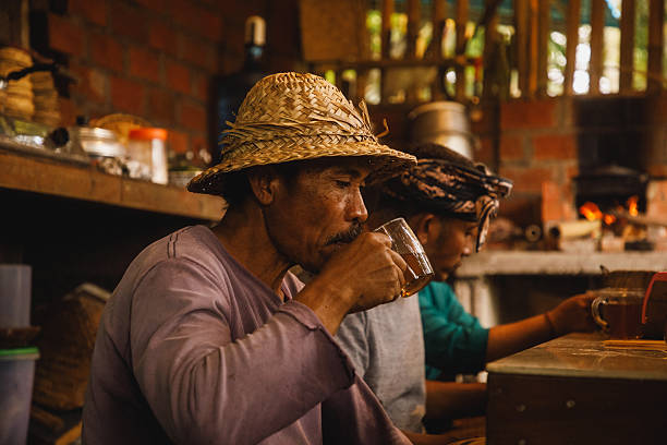 A man wearing a traditional straw hat sips tea in a cozy kitchen with a rustic design. The image captures the warmth of traditional kitchens, featuring wooden interiors and a relaxed atmosphere. The man appears thoughtful and calm, highlighting serene daily life moments.