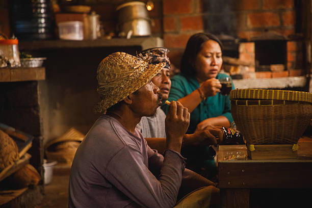 Three individuals gather around a table in a rustic kitchen, indulging in snacks and drinks. They are seated together, emphasizing companionship and culture in a warm atmosphere. Their attire and setting reflect a rural lifestyle, with traditional elements adding authenticity to the scene.