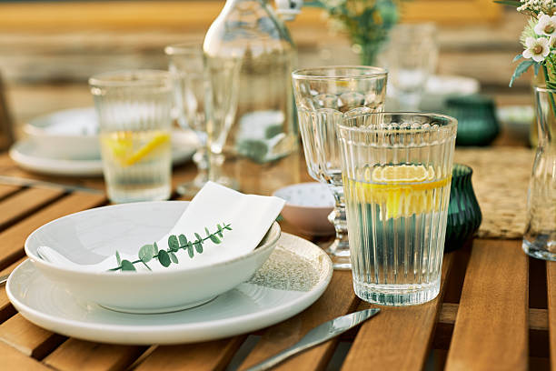 A close-up of a rustic outdoor lunch setting featuring lemon water, leaves, and fresh flowers on a wooden table in natural light.
