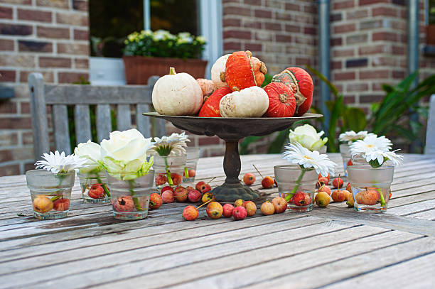 Rustic autumn-themed table decoration with vibrant pumpkins on a pedestal and fresh flowers on a wooden outdoor table, evoking the warmth of the fall season in a cozy setting.