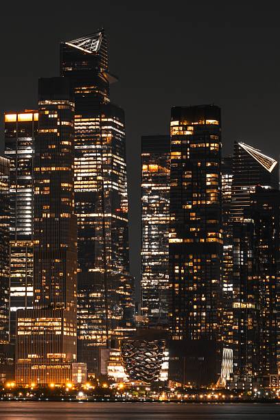 Night view of illuminated skyscrapers in New York City with reflections on the water, showcasing urban architecture and city lights.