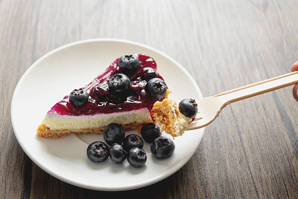 Blueberry cheesecake and fresh blueberries fruit in plate on wooden table. Hand holding fork ready to eat sweet dessert. Fresh blueberry fruit as snack. People enjoy eating blueberries.