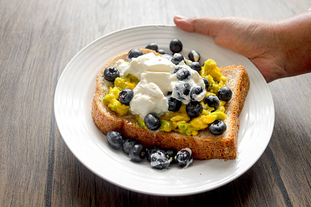 Whole wheat bread with avocado, yogurt and fresh blueberries on top in plate with hand on wooden table. Hand holding toast breakfast ready to eat a meal. Blueberry with toast as a healthy breakfast.