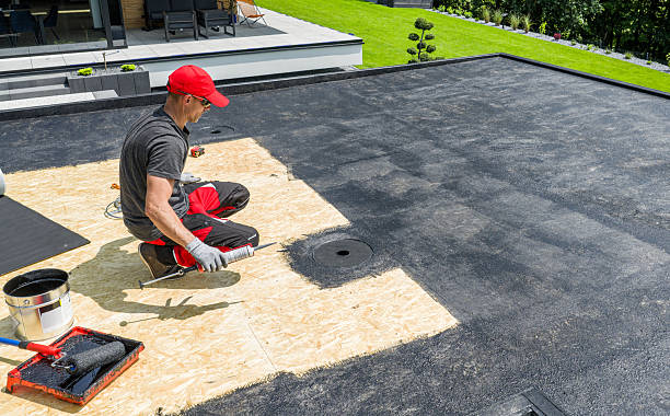 A skilled worker applies roofing material on a flat roof, ensuring proper installation and covering. The residential area features a well-maintained lawn and patio.