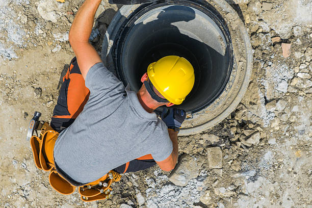 A construction worker in a yellow hard hat repairs a sewer line in an urban construction zone during daylight. Surrounding debris indicates ongoing work.