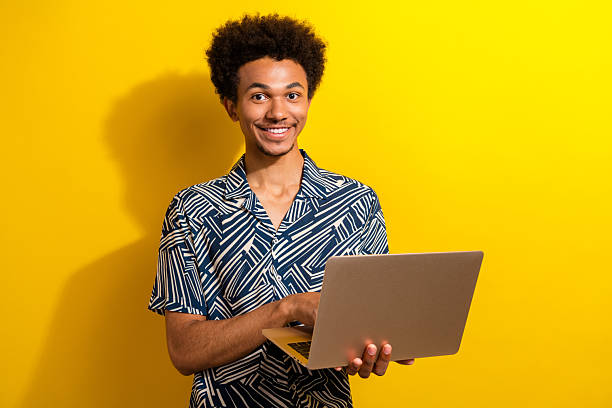 istock Smiling young man with a laptop in a patterned shirt against a vibrant yellow background 2226439450