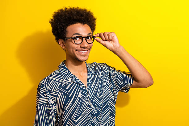 istock Cheerful young man in a patterned shirt adjusts glasses against vibrant yellow backdrop, expressing fun and joy 2226439944