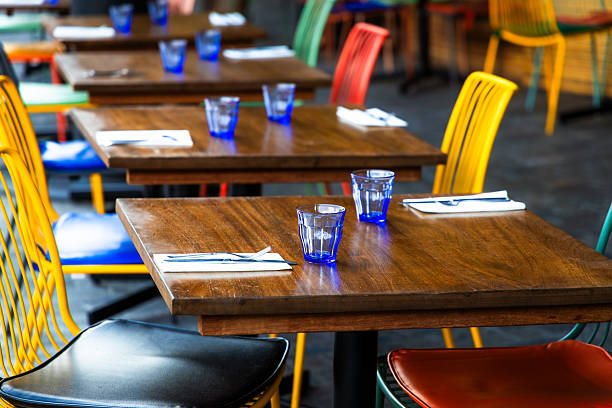 Close up image depicting restaurant tables set with cutlery and glasses and colorful chairs. Close up image with room for copy space.
