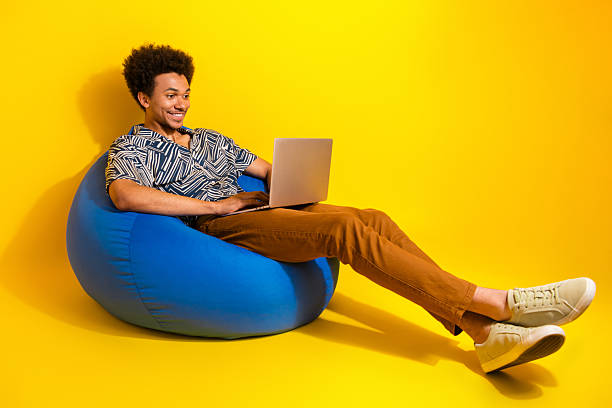 istock Young relaxed man sitting on a bean bag using a laptop on a yellow background embodying leisure and lifestyle 2226440348