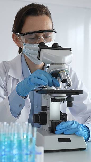 istock Female scientist examining microscope slide, wearing protective gear in laboratory, researching pharmaceutical breakthrough with blue tinted test tube specimens 2226443248