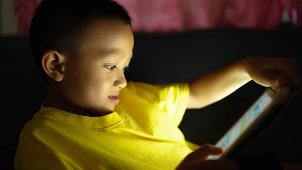 istock Young boy in bright yellow shirt is intently focused on tablet screen, illuminated by device light. soft glow highlights his concentration and curiosity, creating warm and engaging atmosphere 2226451977
