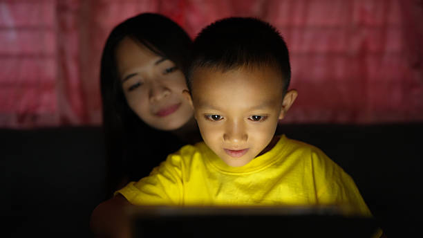 A child in yellow shirt is focused on tablet screen, illuminated by its light, while woman sits behind, watching with gentle smile. scene captures warm moment of connection and curiosity