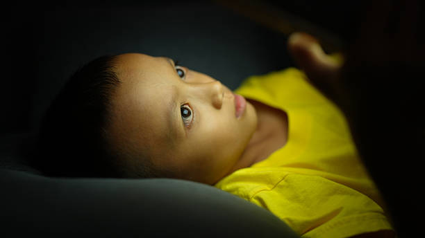 A young child with short hair is lying down, gazing intently at light source, wearing bright yellow shirt. soft lighting creates warm and focused atmosphere