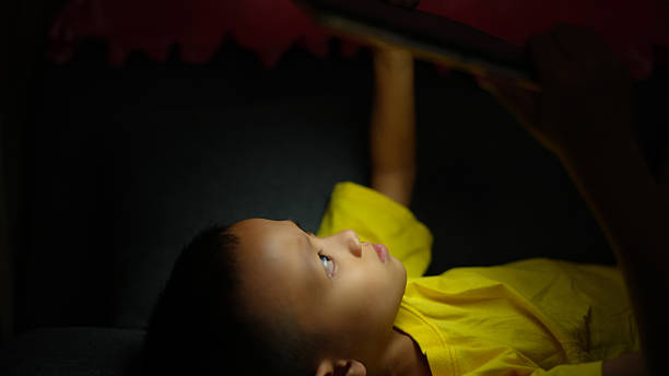 istock Child in yellow shirt is lying on dark surface, gazing intently at tablet. soft light highlights their focused expression, creating serene atmosphere 2226452090
