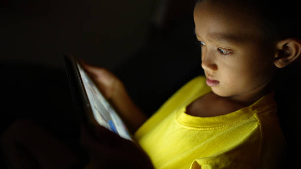 Young child is focused tablet screen, illuminated by its light, wearing bright yellow shirt. scene captures moment of concentration and curiosity, highlighting interaction between technology