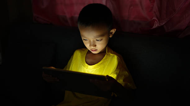 istock Young child in yellow shirt is intently focused on tablet screen, illuminated by its glow in dimly lit room, creating captivating atmosphere of concentration and curiosity 2226452111