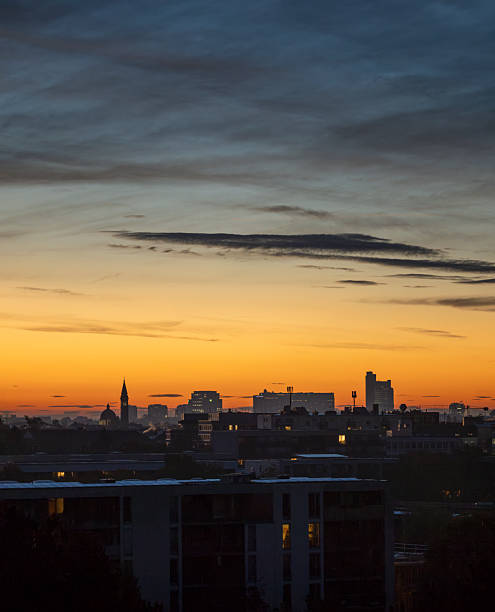 A closer look at the east side of Munich at dawn, with silhouetted buildings, church towers, and office lights under a golden-orange sky.