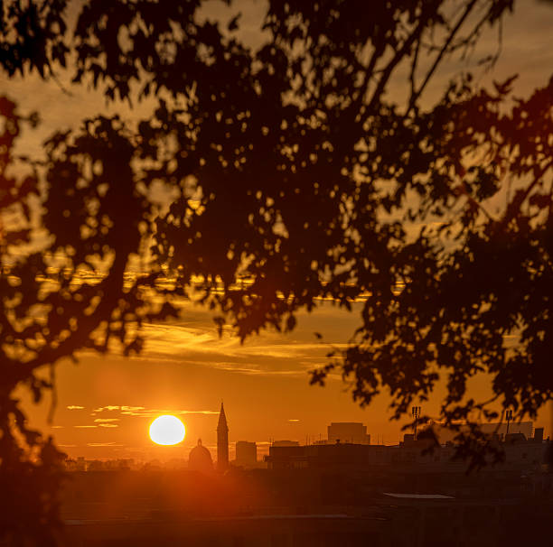 A vibrant sunrise rises above the Munich skyline, framed by silhouetted leaves and urban rooftops. Warm golden light highlights the city’s iconic church spire.