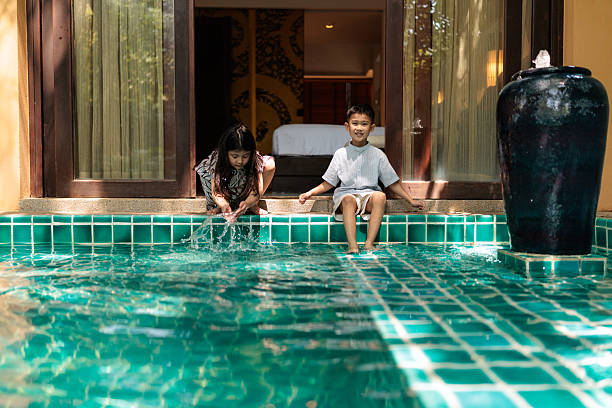 Two joyful children enjoying playful moments by the villa pool, the boy sits on the edge, dipping his feet into the cool water, while the girl happily splashes and plays with her hands in the water. Their laughter and smiles capture the carefree fun of a family vacation in a beautiful tropical setting.