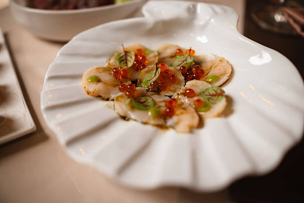 Seared scallops seasoned with tobiko roe, microgreens, and a drizzle of sauce, served on a seashell-shaped plate in a restaurant.