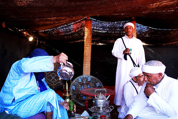 Man dressed in beautiful berber clothings making mint tea for his guests inside a traditional jaima tent.\nThe Berber tea ceremony is more than just making and drinking tea. It represents a rich social tradition, a gesture of hospitality, and a way to build personal relationships. \n\nThis is part of the celebrations of cultures and lifestyles of the desert of the Festival Moussem of Tan Tan that takes place every spring in the southern city of Morocco.