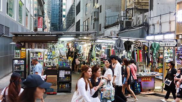 Causeway Bay, Hong Kong- 8 Jun 2025: a bustling alleyway with modern skyscrapers and a graffiti-covered building. Covered tin shed selling grocery, umbrella and clothes with busy crowd walking
