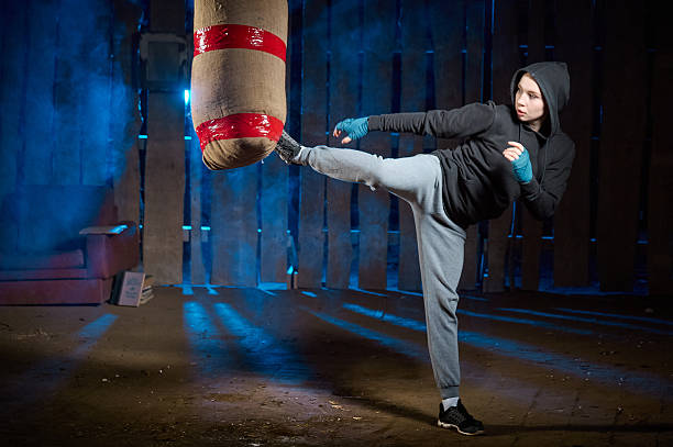 Cute teen girl dressed a black hoodie and white boxing gloves on black and dark-blue background. The concentrated girl is boxing blows to a makeshift punching bag. Shooting in smoke at an abandoned building