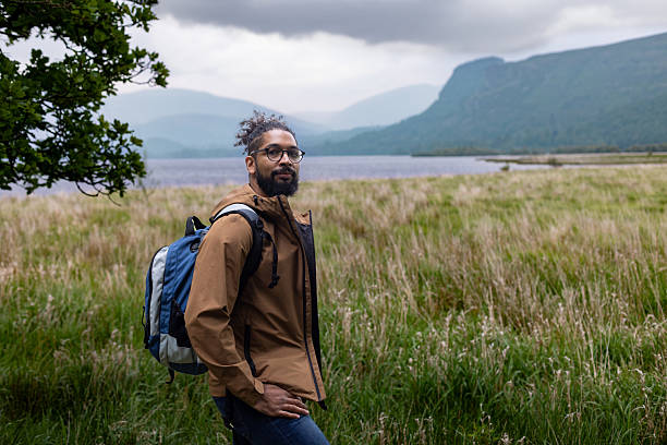 A wide portrait shot of a man wearing a brown waterproof jacket and backpack, standing beside a tree in the countryside near Keswick in the Lake District, UK. Mountain peaks and tall grass stretch into the distance under an overcast sky, capturing the calm beauty of a rural hiking adventure. Derwent Water can be seen behind him.Videos are available similar to this scenario.