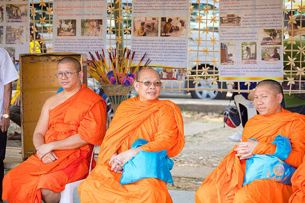 Group of thai monks in open public ceremony captured in and at Kelang Nakhon Public Park in Lampang city. Lampang is in north Thailand. In scene three mons are siting on chairs. Two monks are wearing eyewear. Two also have blue bags on their legs. One is looking towards another one. In background are information billboards with photos and texts about thai craft on making traditional large thai drums. A fouth man is standing at left side and is partly visible.