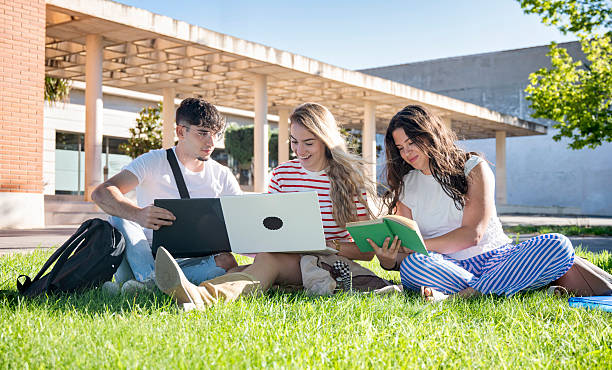 istock Happy university students sitting on green campus lawn smiling and studying together with laptop and book on a sunny day 2226493040