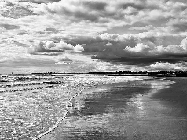 A tranquil sunrise scene at Cocoa Beach, Florida, featuring gentle ocean waves rolling onto a wide sandy shoreline under a dramatic, cloud-filled sky. The early morning light creates soft reflections on the wet sand while textured clouds add depth and atmosphere to the coastal landscape.