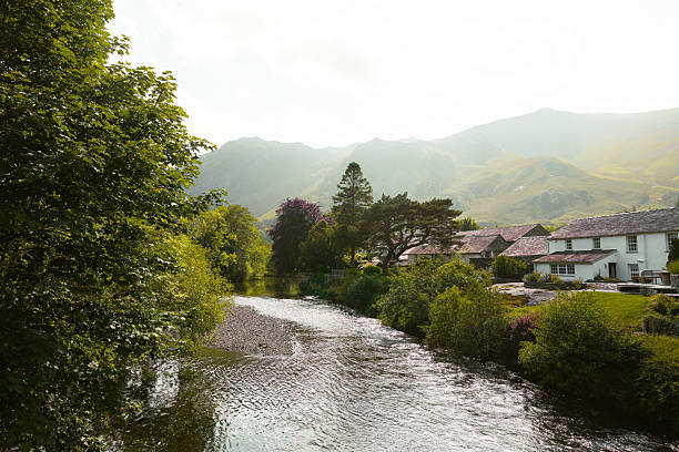 A wide angle view of a flowing lake in Borrowdale in Cumbria in the Lake District, The lake is lined by luxuriant green abundant blooming trees and the late afternoon misty sky gnetly blurs the mountain peaks in the distance.
