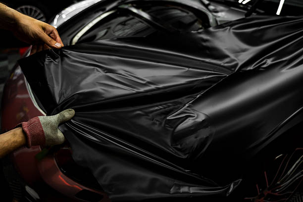 istock Car wrapping process using black matte vinyl stretched into place. Careful application and tensioning of black vinyl avoids surface damage or bubbling. Matte black vinyl is applied across car hood. 2226498345