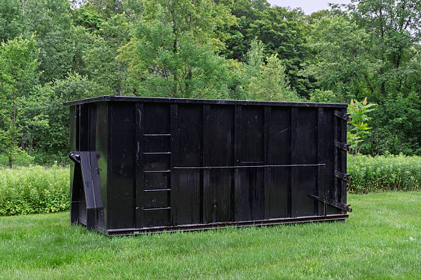 A large industrial black roll-off dumpster placed on a grassy lawn with lush green trees behind. Suitable for concepts like construction waste, home renovation cleanup, junk removal, and rural property maintenance.