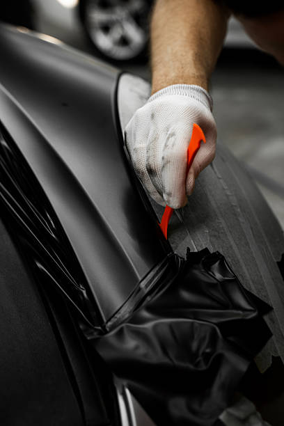 istock Trim tool to apply matte black wrap around a car panel edge. Car Wrapping with Trim Tool. Car detailing applies vinyl wrap around trim using plastic wedge tool for seamless finish. 2226498884