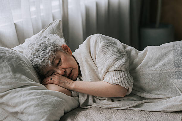 A senior Japanese woman with gray curly hair sleeps peacefully on her side in bed during the day. She appears calm and relaxed in a soft, cozy home environment, dressed in light knitwear, with natural daylight filtering through the curtains. Concept of healthy aging, rest, relaxation, wellness, and peaceful lifestyle.