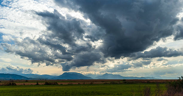 Cloud formations dominate this scenic landscape featuring a flat green field and distant mountains under an overcast sky. This stunning natural view highlights atmospheric contrasts and inspires awe, tranquility, and wonder, making it ideal for themes involving nature's raw beauty.