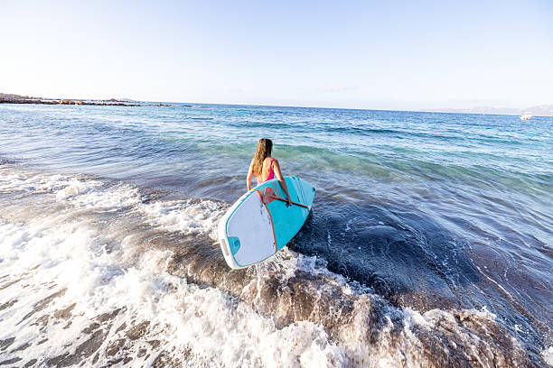 Playa de Las Americas, Tenerife, Canary Islands, Spain - June 3, 2020: young male surfer wearing a wetsuit and holding a large surfboard while walking the connecting promenade and looking at the ocean.