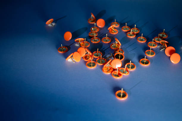 Orange push pins scattered on a smooth, blue surface. The pins are circular with metallic tips, predominantly facing upward, suggesting disarray. The vibrant contrast between the orange and blue enhances the visibility of the pins, making them stand out. The scene is well-lit, highlighting the glossy texture of the pins and casting subtle shadows on the surface, conveying a casual yet organized atmosphere. Suitable for themes of organization or office supplies.