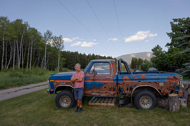 Senior man leans against rusty pickup truck in alleyway near forest, Crowsnest Pass, Alberta