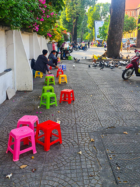 Ho Chi Minh City, Vietnam- March 26, 2025: Locals sit on bright plastic stools along tree-lined sidewalk in Vietnam, with pigeons and scooters nearby